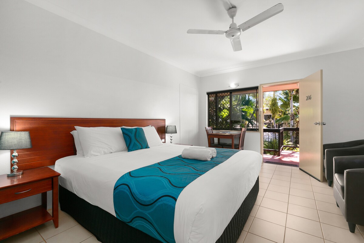 The King Suite features a spacious layout, with a large bed covered in crisp white linens and a decorative teal throw. A door leads to a balcony with views of tropical greenery. A dining table and chairs are positioned near the entrance, alongside two armchairs.