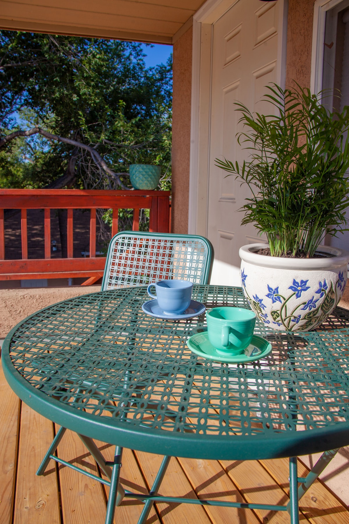 A round, turquoise metal table is set on a wooden deck, accompanied by two chairs. A blue cup and a green cup sit on a matching tray, with a potted plant featuring vibrant flowers positioned nearby.
