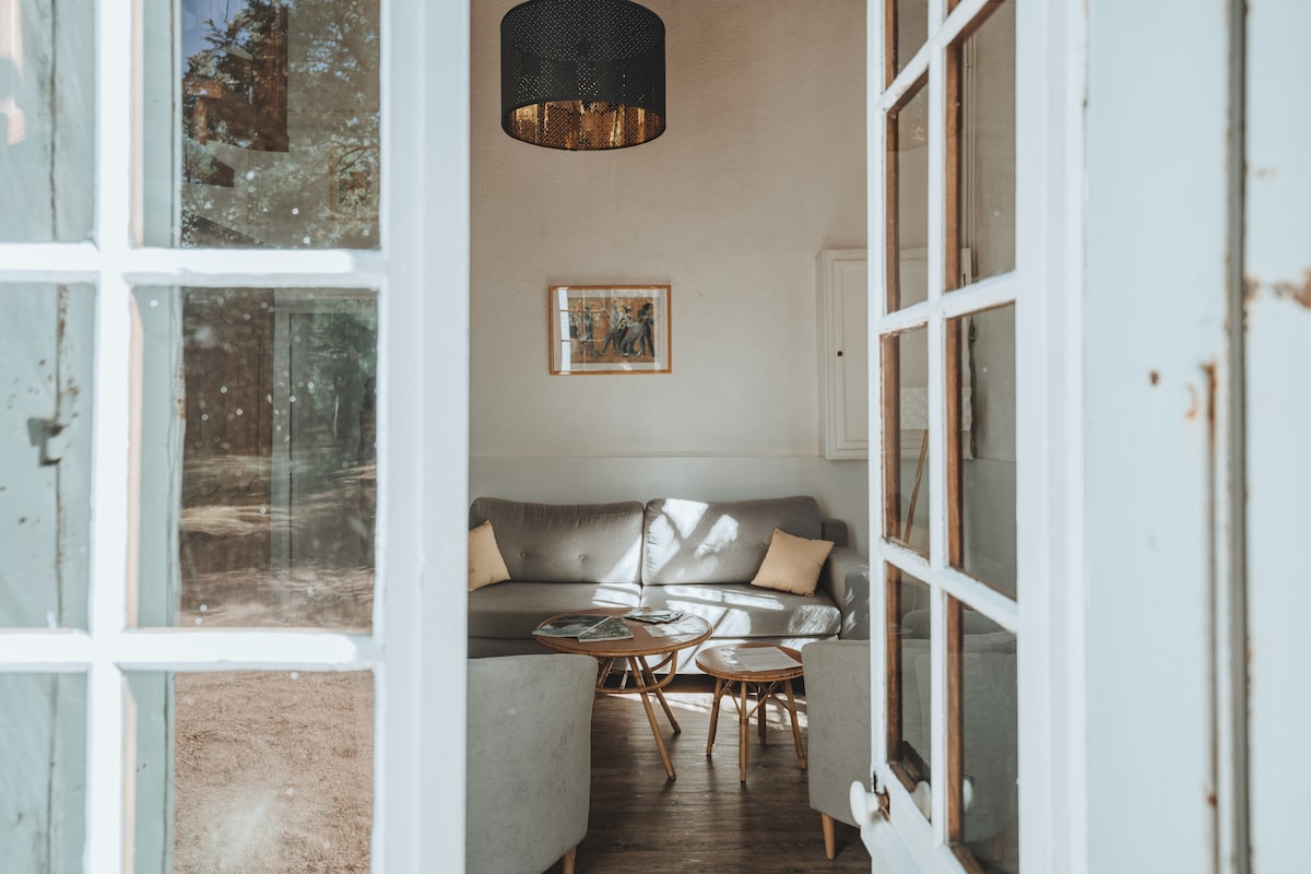 The inviting lounge is visible through an open glass door, featuring a modern gray sectional sofa accompanied by two light-colored cushions. A round coffee table sits at the center, topped with magazines. A warm overhead light enhances the calm atmosphere.