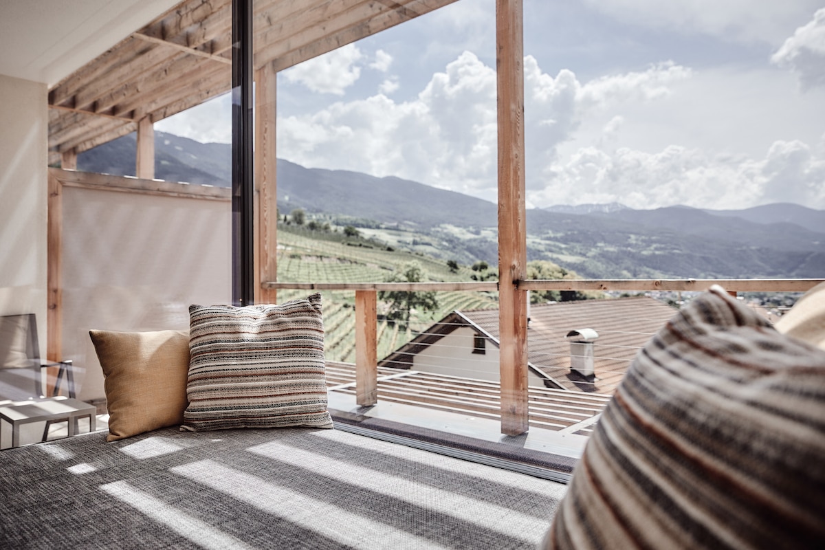 A comfortable seating area features large pillows and a soft rug, positioned near expansive windows. The stunning view includes rolling vineyards and mountains under a partly cloudy sky, allowing natural light to fill the space.
