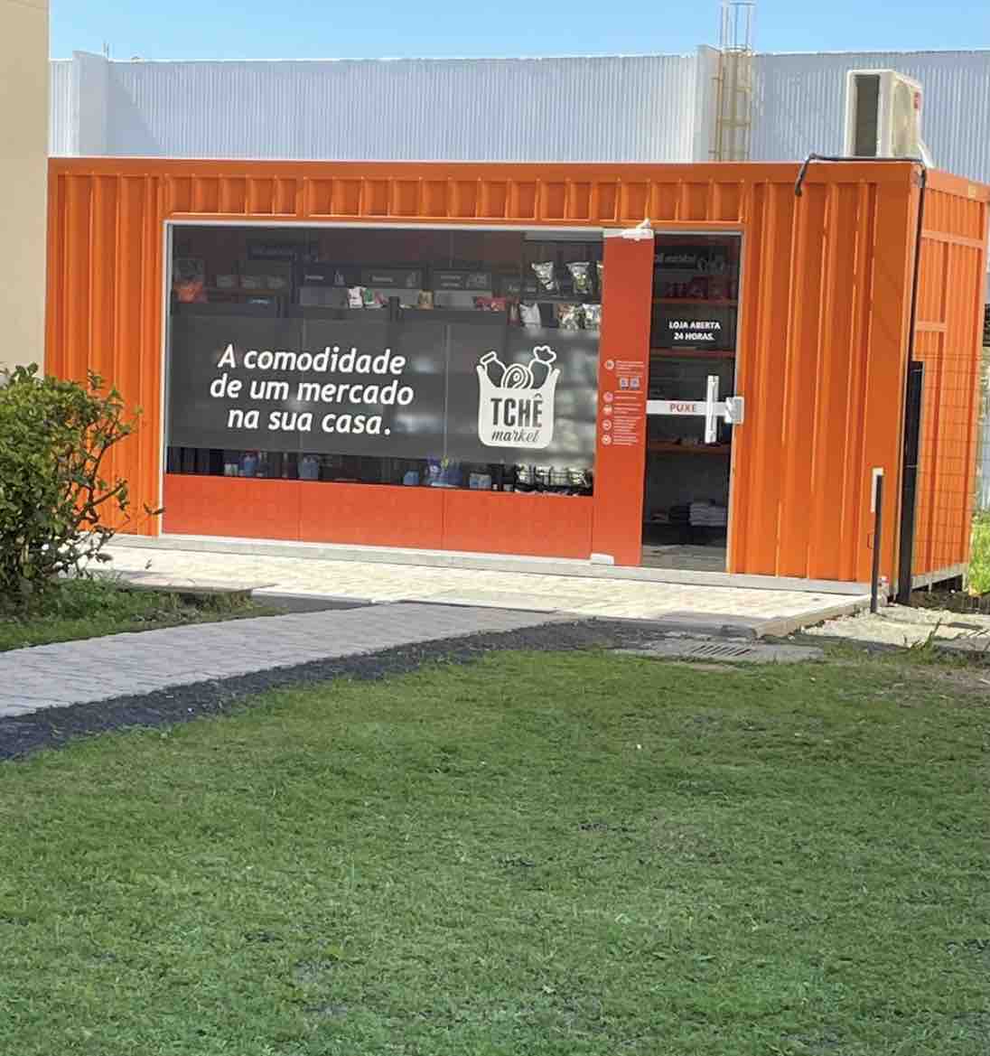 An inviting market entrance is marked by a bright orange facade. The large window displays various goods available inside, with a sign that translates to 'The convenience of a market at your home.' A well-kept grassy area leads up to the entrance.