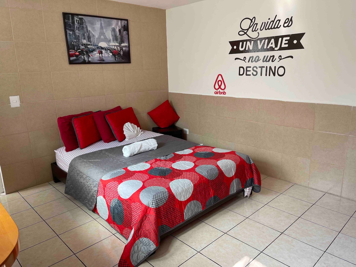 A spacious sleeping area features a large bed covered with a colorful red, gray, and white quilt. Decorative pillows and a rolled towel are placed at the head of the bed. A wall mural with the phrase 'La vida es un viaje, no un destino' adds character to the room.