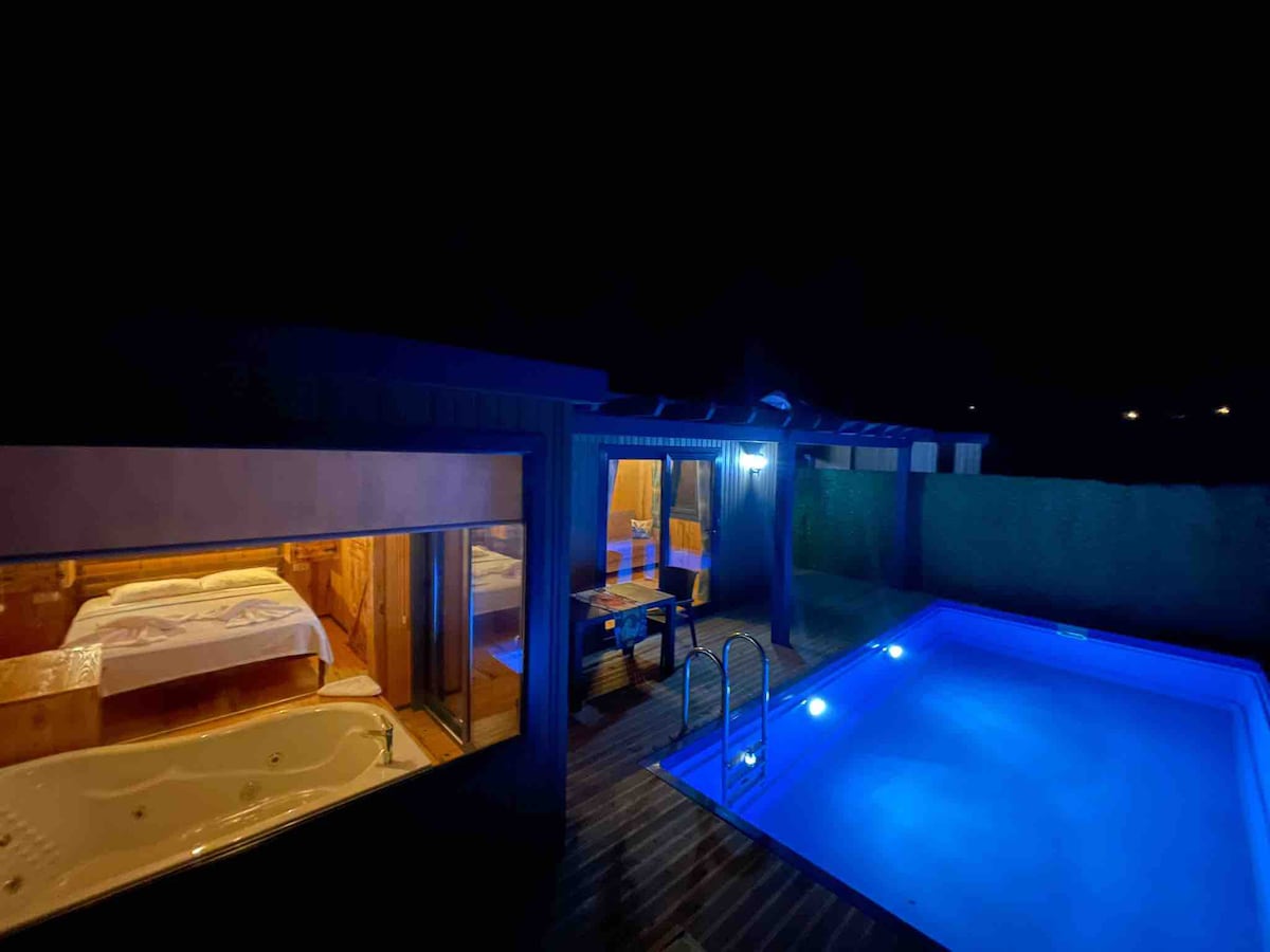 A serene outdoor area features a glowing blue pool, providing a contrast against the night sky. Adjacent to the pool, a spacious indoor space is visible through large glass windows, showcasing a cozy sleeping area and a relaxing jacuzzi.