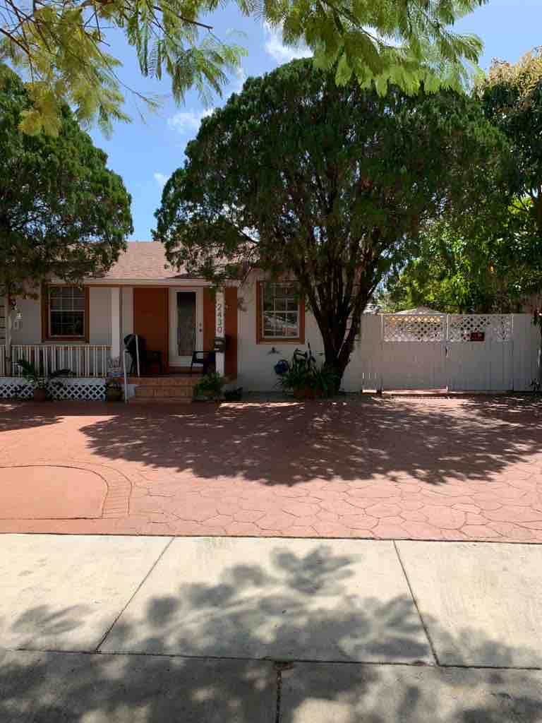 A front view of a charming studio, framed by two large trees that provide ample shade. The exterior features a light-colored façade with an orange door and windows. A paved area leads to the entrance, accompanied by a white picket fence and greenery around the surrounding space.
