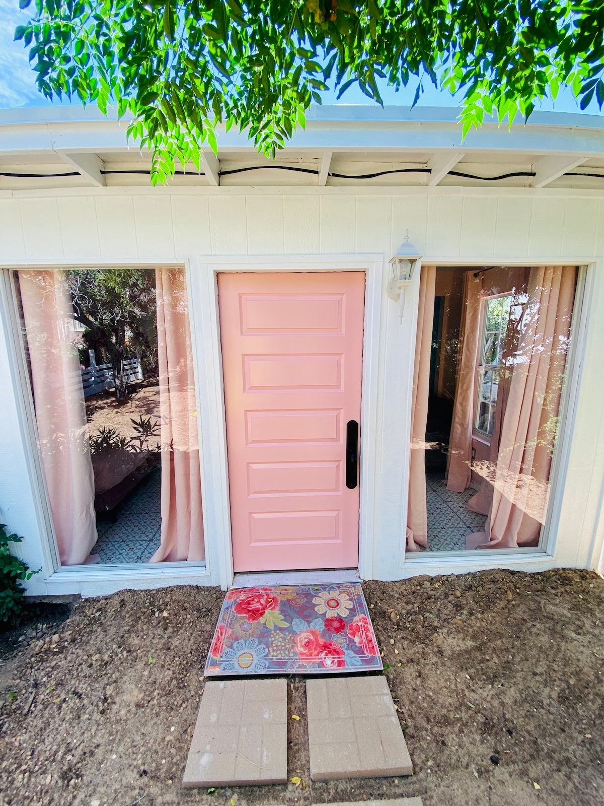 A charming entrance is presented with a pink door surrounded by large windows adorned with soft, light-colored curtains. Floral mats on the ground lead toward the entrance, while greenery from nearby plants provides a peaceful touch to the space.