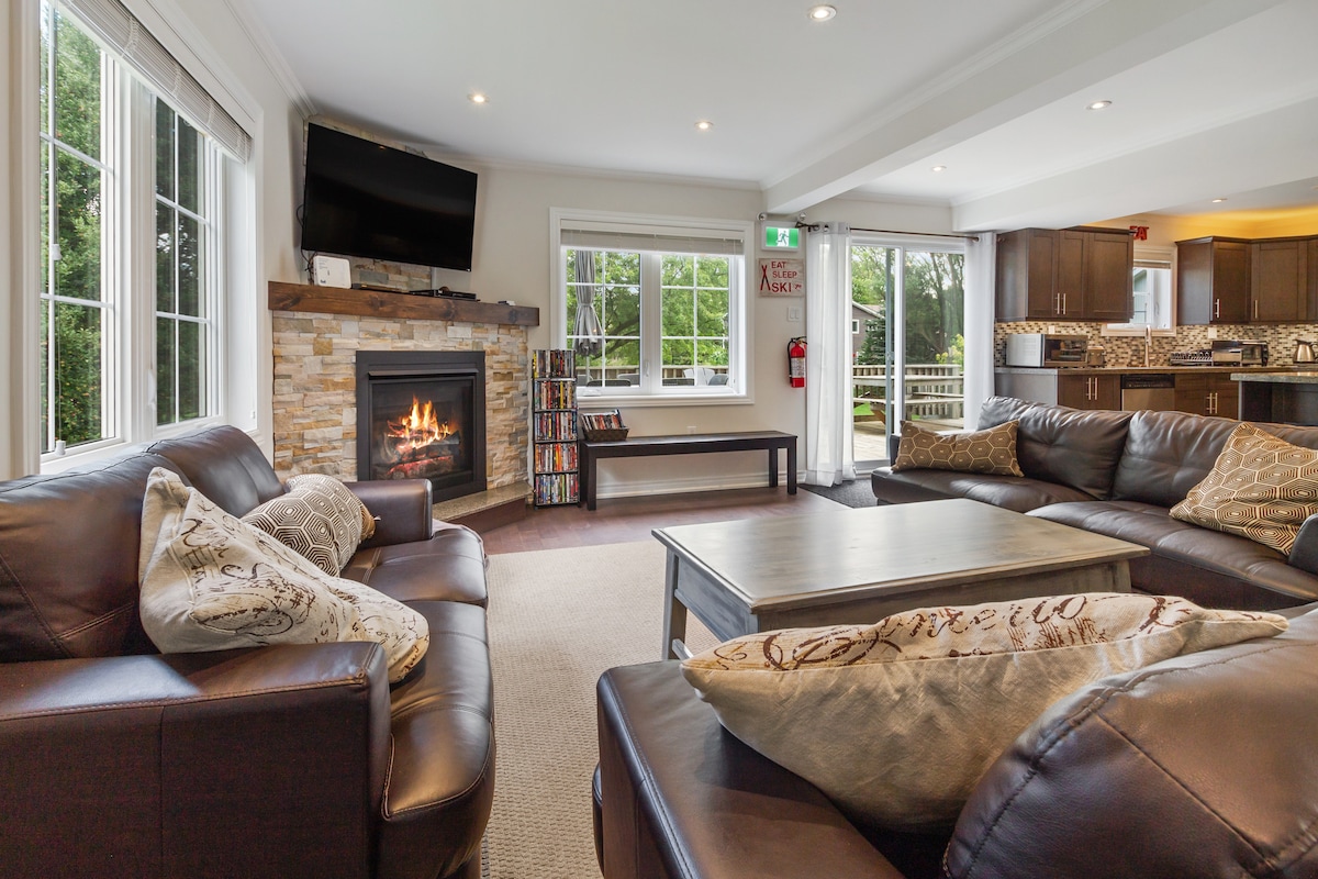A spacious living area features a large sectional sofa with decorative pillows, centered around a gas fireplace encased in stone. Natural light floods the room through multiple windows, and a flat-screen TV is mounted above the fireplace. The open layout connects to the kitchen area in the background.