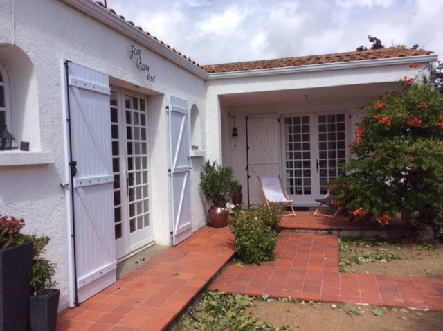 A charming exterior features whitewashed walls and tiled roof accents. The entrance is framed by wooden doors, leading to a cozy patio. Potted plants and vibrant foliage enhance the inviting space, with a sun lounger positioned for relaxation among well-kept gardens.