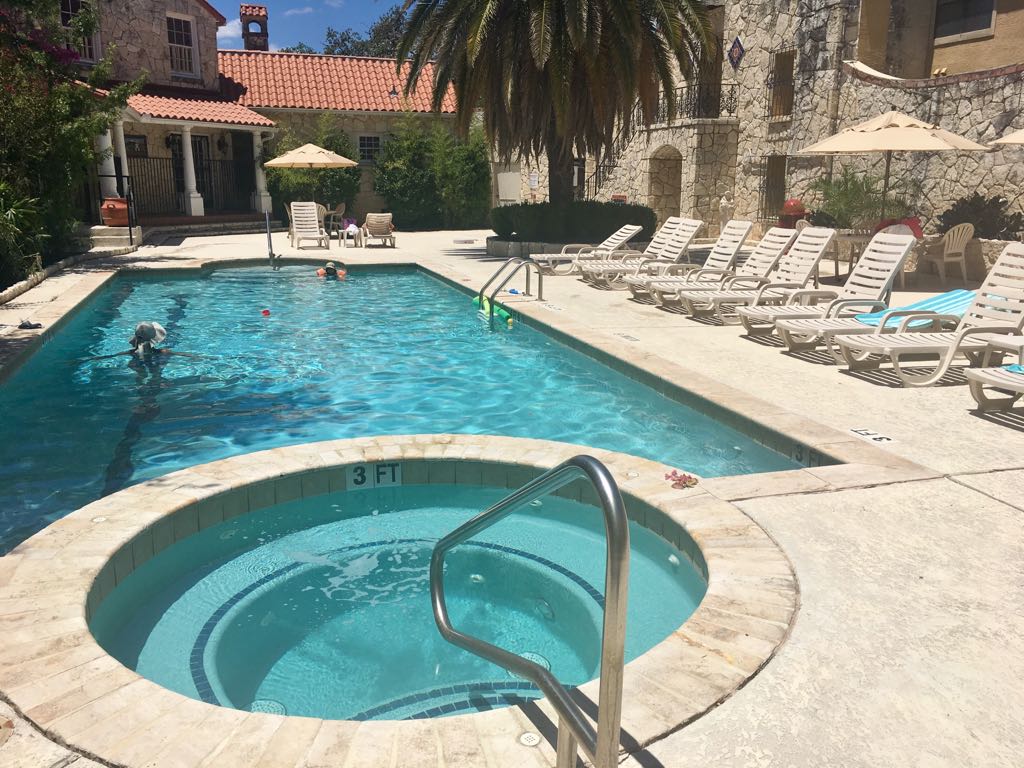 A clear swimming pool is surrounded by a large patio area featuring multiple lounge chairs and umbrellas. A hot tub is positioned adjacent to the pool, with stairs leading into the water. Lush greenery is visible in the background, adding to the serene environment.