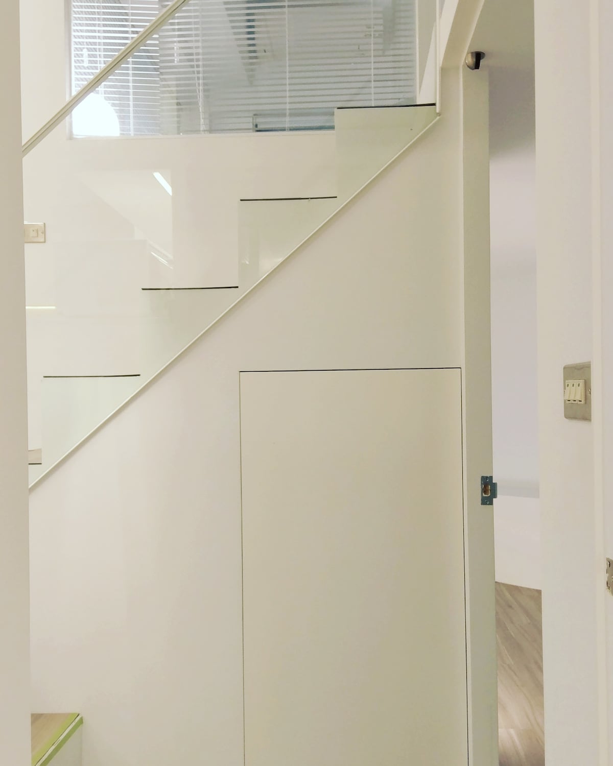 A staircase with glass railings creates a sleek, modern aesthetic. The staircase is situated against a white wall, enhancing the minimalist design. A small closet door is visible at the base of the stairs, maintaining the streamlined look of the space.