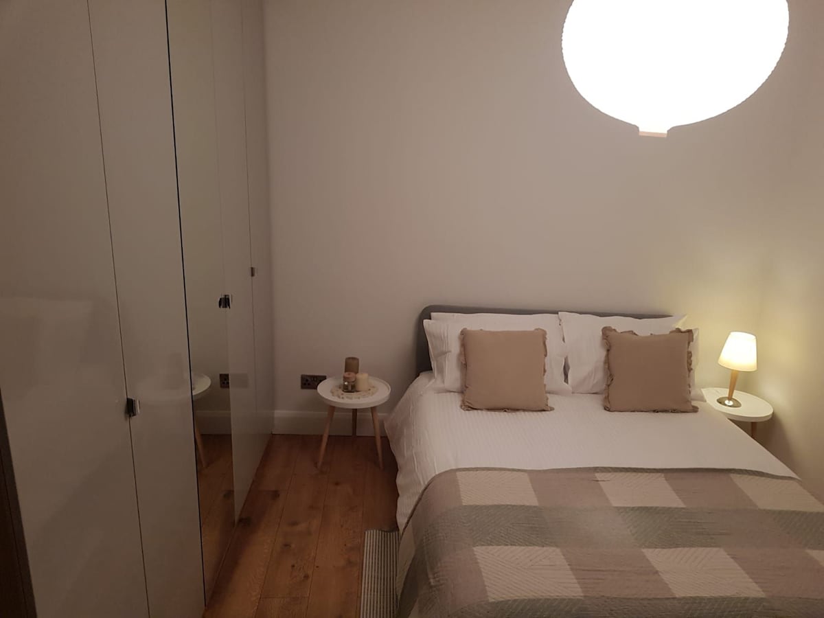 A cozy sleeping area features a double bed with neutral-toned pillows and a light blanket. A bedside lamp provides soft lighting, and a minimalistic side table is positioned next to the bed. A wardrobe with reflective doors adds depth to the room, complemented by warm wooden flooring.