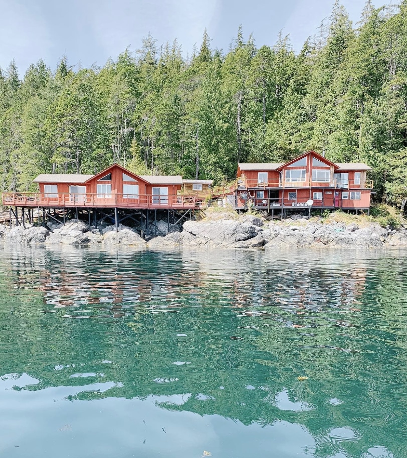 Two red cabins are nestled on rocky waterfront, surrounded by lush green trees. Their large windows reflect the tranquil water, creating a seamless connection between the cabins and the natural landscape. The calm, clear water enhances the serene setting.