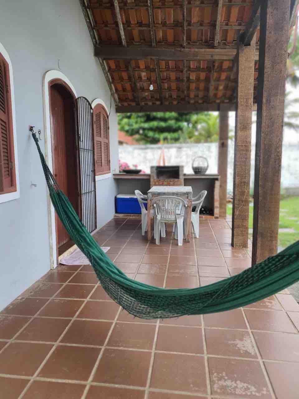 A covered patio area is visible, featuring a green hammock hanging on one side. A rectangular table with four white plastic chairs occupies the center. An outdoor grilling area can be seen in the background, surrounded by greenery and a serene atmosphere.