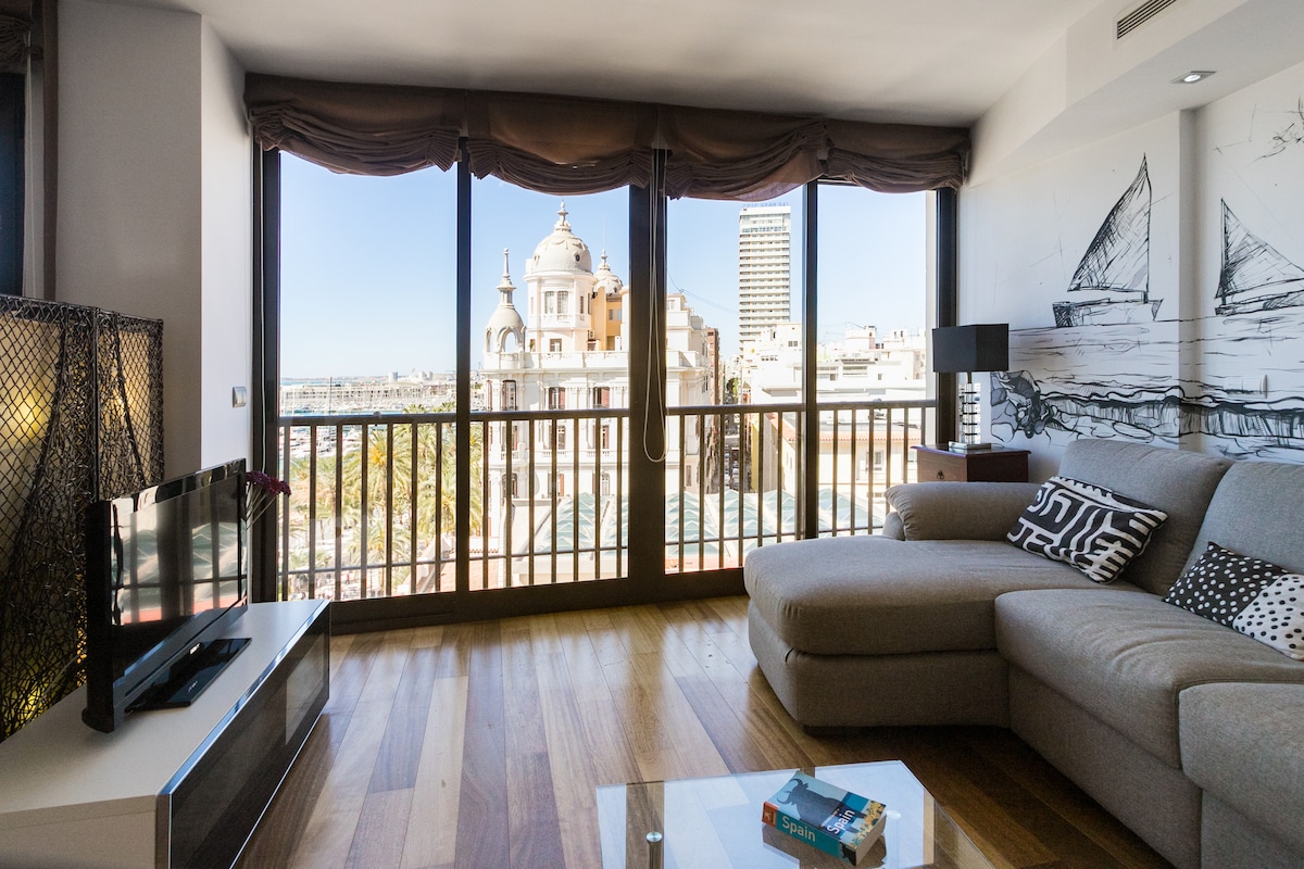 A bright and airy living space features large windows offering views of the cityscape. A comfortable gray sectional sofa is positioned near a wooden coffee table, alongside a television on a low media unit. Light wood flooring complements the modern decor.