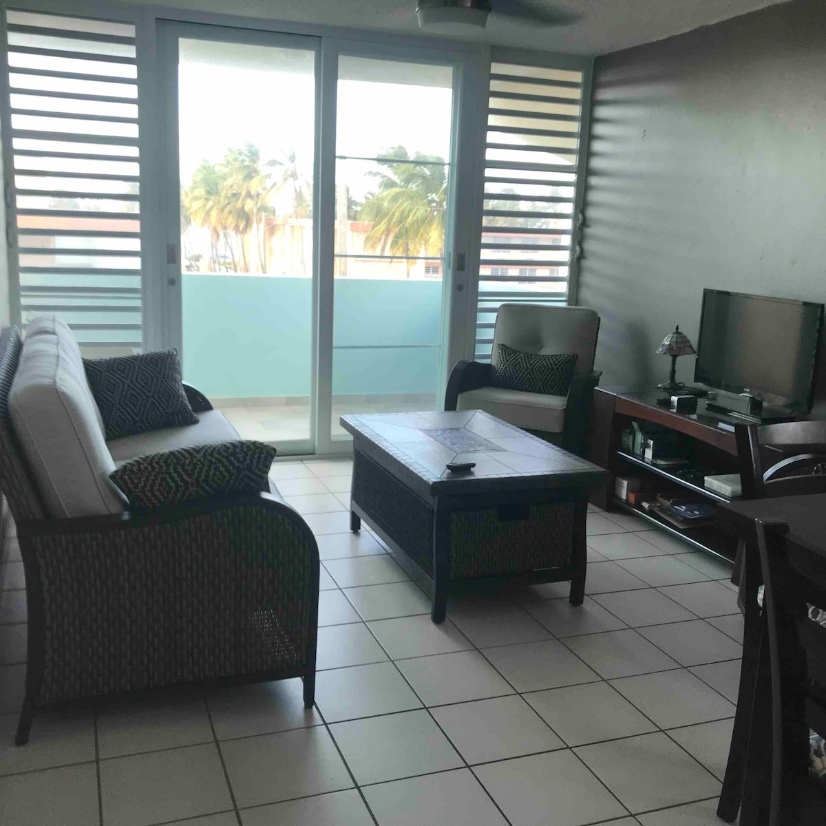 A cozy living area is filled with natural light from large sliding glass doors that open to a balcony. Comfortable seating includes a sofa and a chair, complemented by a coffee table. A television is situated on a dark wood console against a grey wall.
