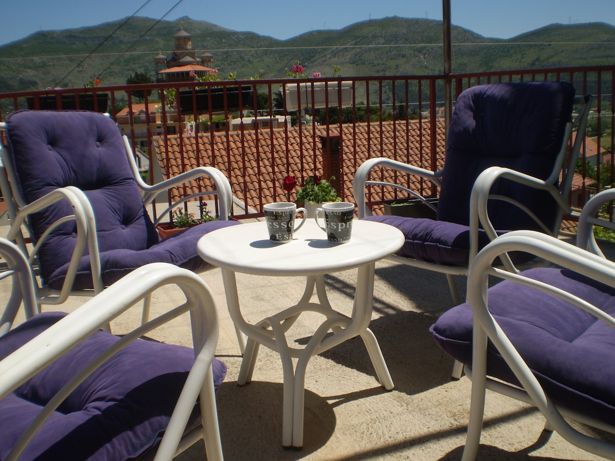 A cozy outdoor seating area is set up with four lounge chairs featuring plush purple cushions. A small table sits in the center, with two cups placed on it. The surrounding view includes rolling hills and greenery, complemented by potted plants along the railing.