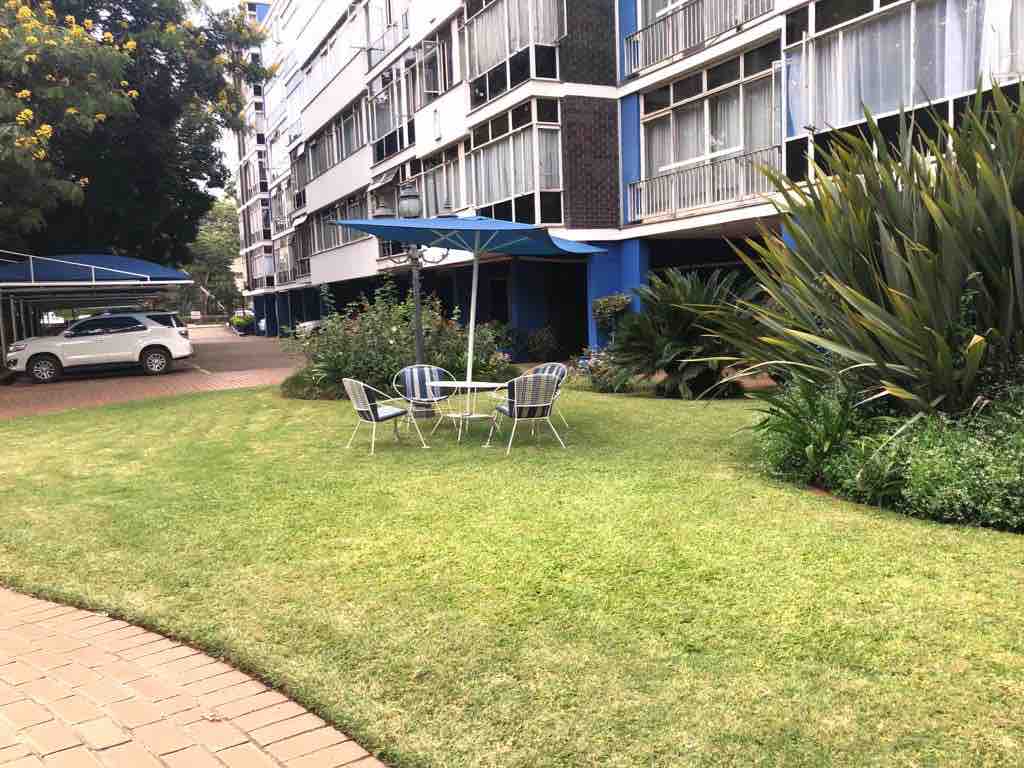 A neat garden area is visible, featuring well-maintained grass and flower beds. A small table with four chairs is situated under a blue canopy, providing a shaded outdoor seating option. Surrounding greenery enhances the calming atmosphere, set within a residential complex.