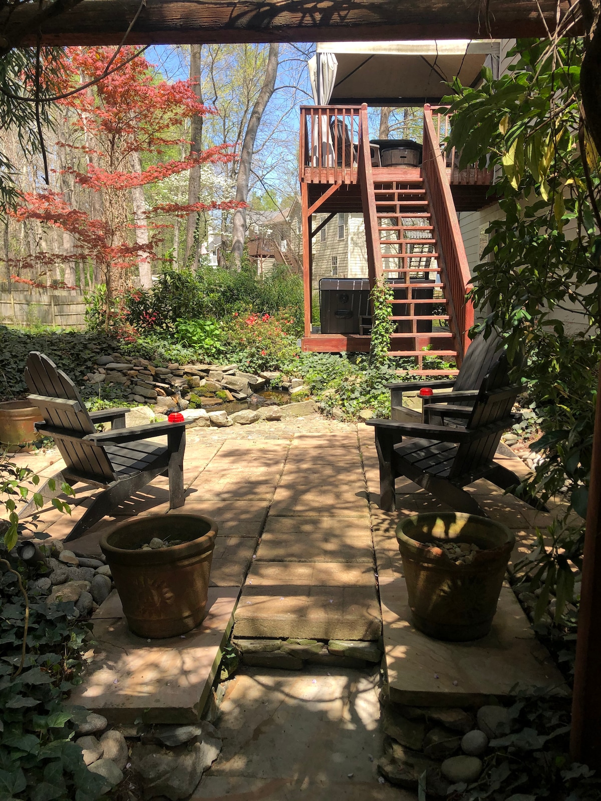 A private garden area is displayed, featuring a stone path leading to two wooden chairs. Potted plants enhance the natural ambiance, while greenery envelops the space. A deck with steps is visible in the background, providing access to the upper level.