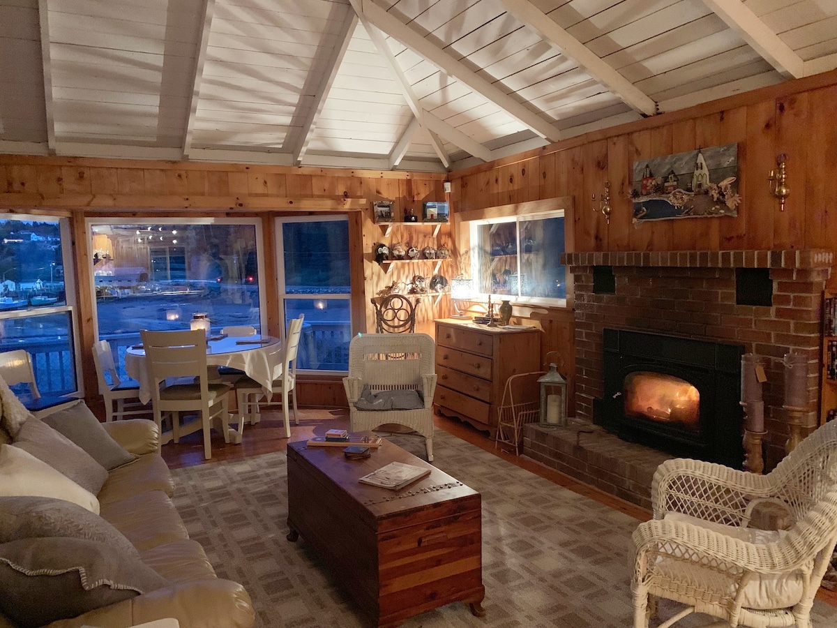 A cozy living area features wooden walls and a high ceiling with exposed beams. A comfortable sofa and wicker chairs surround a wooden coffee table. A warm glow emanates from the brick fireplace, while large windows provide views of the harbour and nearby landscape.