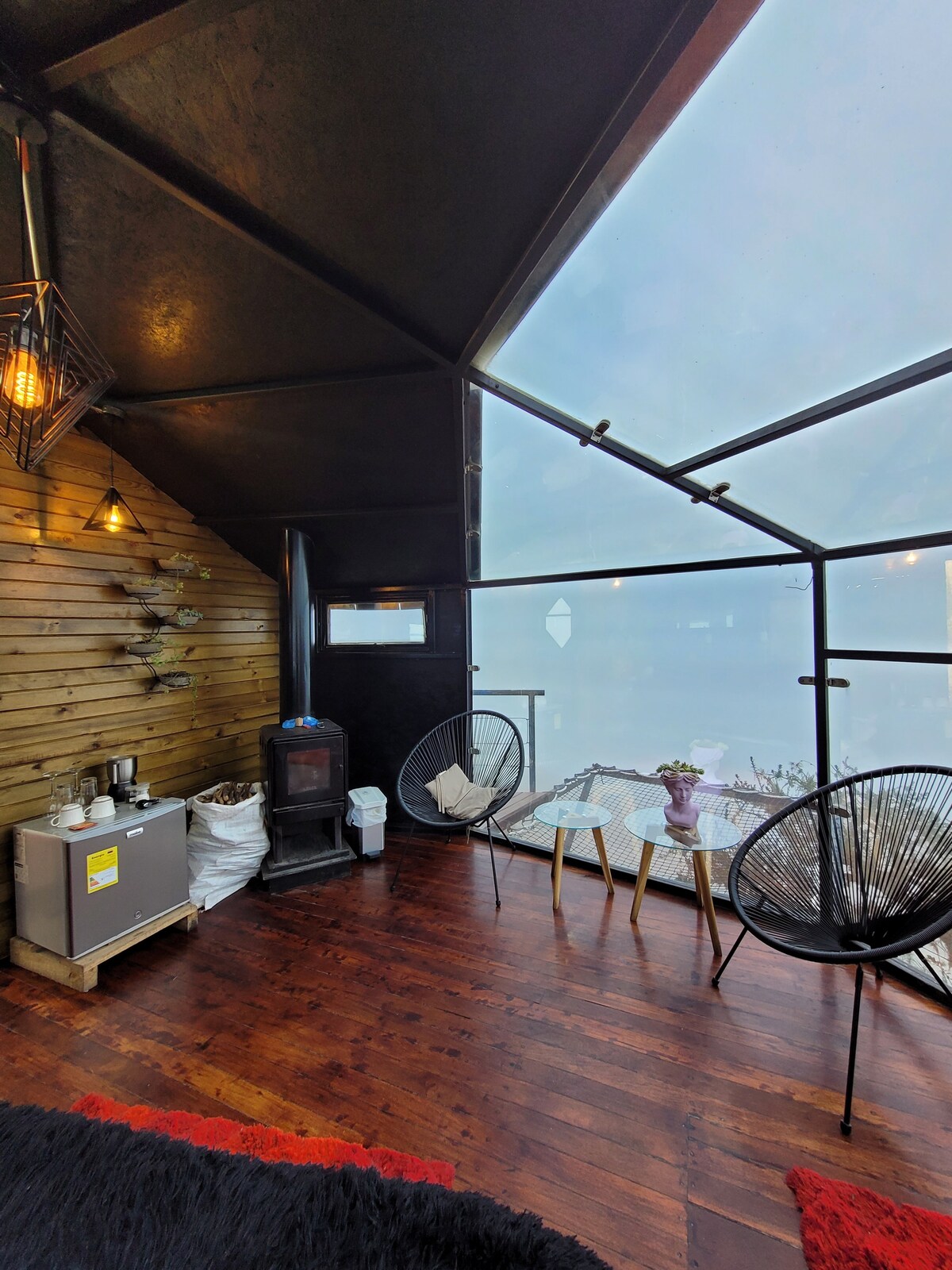 A cozy seating area features two Acapulco chairs positioned around a small glass table. A wooden wall adds warmth, while a sleek fireplace and a minibar are located in the corner. Large glass panels provide a clear view of the surroundings.