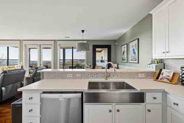 A modern kitchen features sleek white cabinetry and stainless steel appliances, including a refrigerator and dishwasher. The countertop is finished with a subtle grey and white patterned backsplash, while large windows allow natural light to illuminate the space, offering a view of the surrounding area.