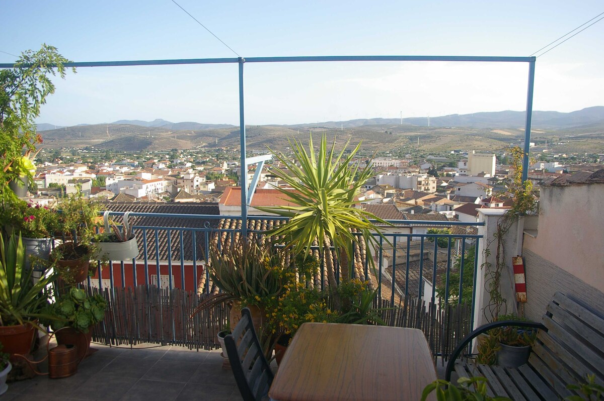 A terrace is featured overlooking a scenic view of the surrounding hills and nearby town. Potted plants add greenery to the space, and a small table accompanied by chairs provides a cozy spot for relaxation. Clear skies enhance the tranquil atmosphere.