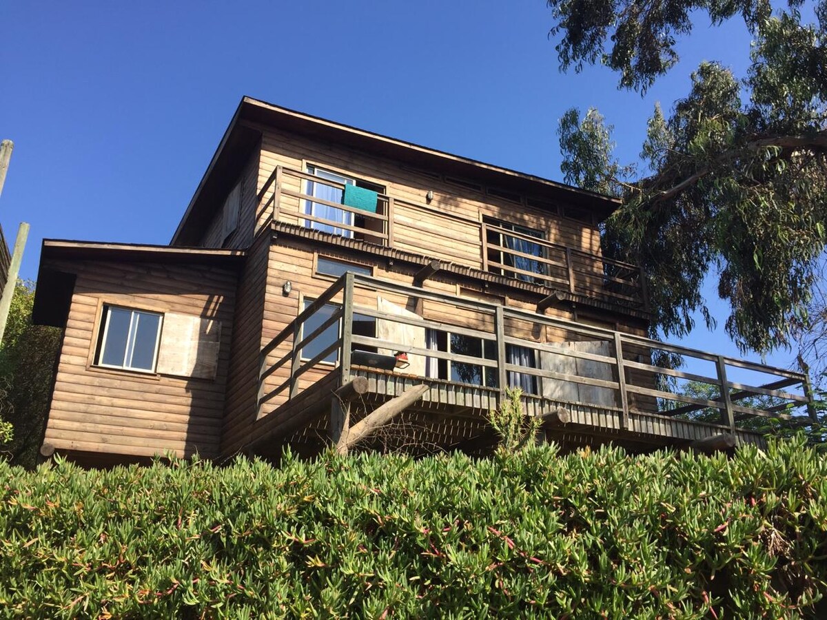The wooden house features two stories with balconies, surrounded by lush greenery. Large windows allow natural light to fill the interior. A wooden deck extends from the first floor, providing access to outdoor areas. The clear blue sky complements the tranquil setting.