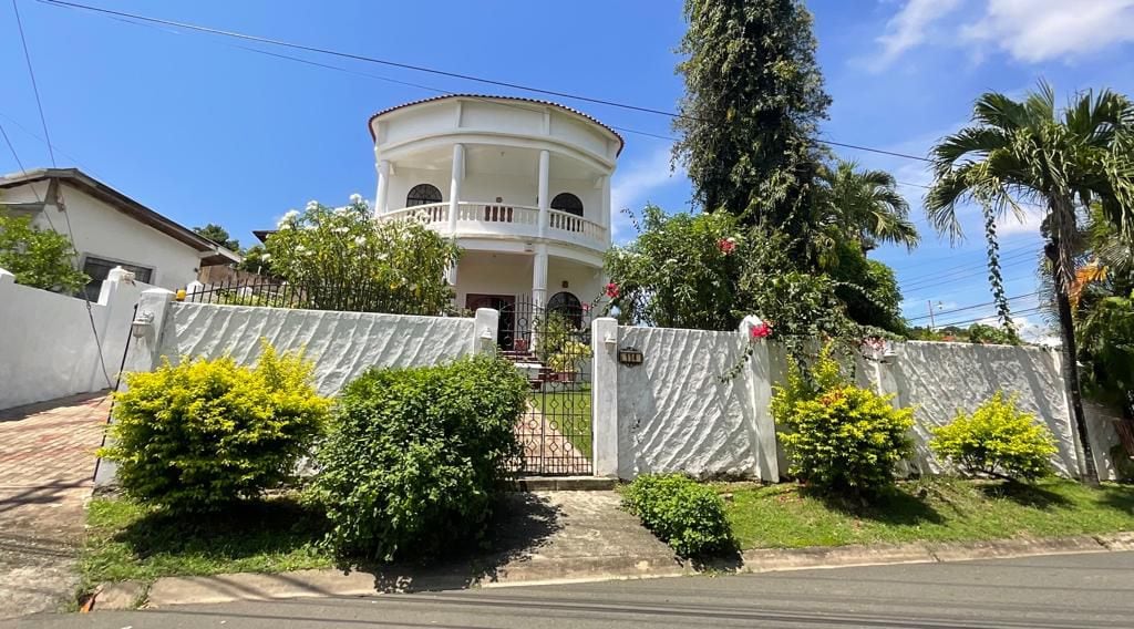 A charming colonial-style villa is visible from the street, featuring a round upper level with a balcony and white railings. Lush greenery and colorful flowers frame the property, complemented by a well-maintained white fence and a pathway leading to the entrance.
