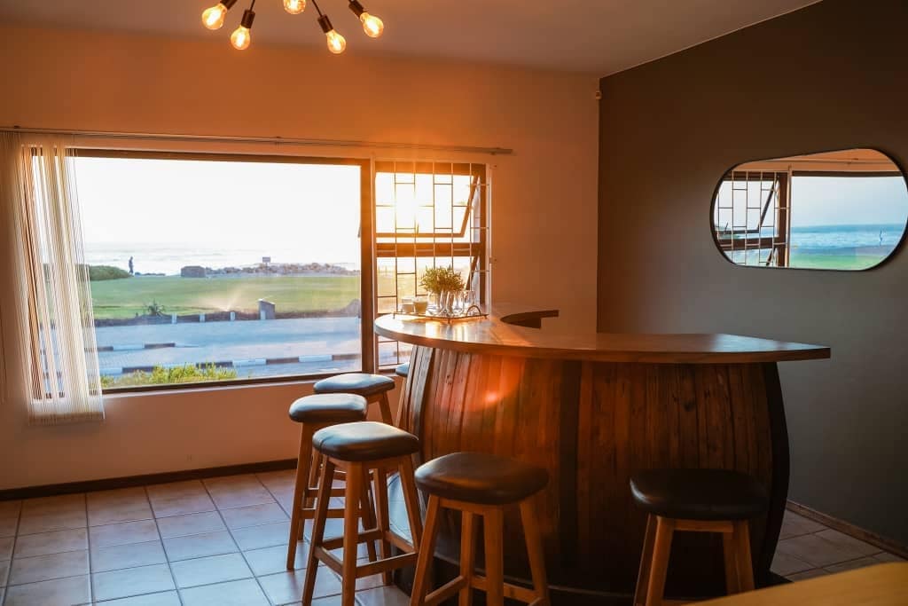 A wooden bar counter is positioned beside a large window, offering views of the sea. Four bar stools are arranged around the counter. Natural light filters through sheer curtains, creating a warm ambiance. A decorative mirror is mounted on the wall opposite the window.