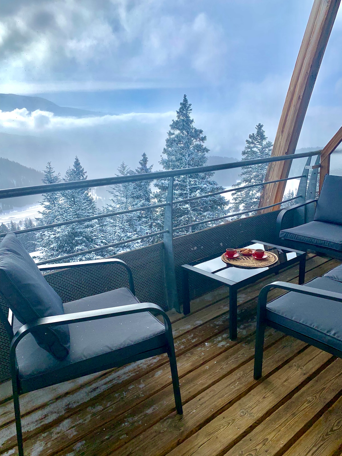 A balcony reveals a serene view of snow-covered mountains and trees. Comfortable seating is provided by two armchairs and a small table. A platter holds fruit, adding a touch of warmth to the cool, misty atmosphere surrounding the outdoor space.
