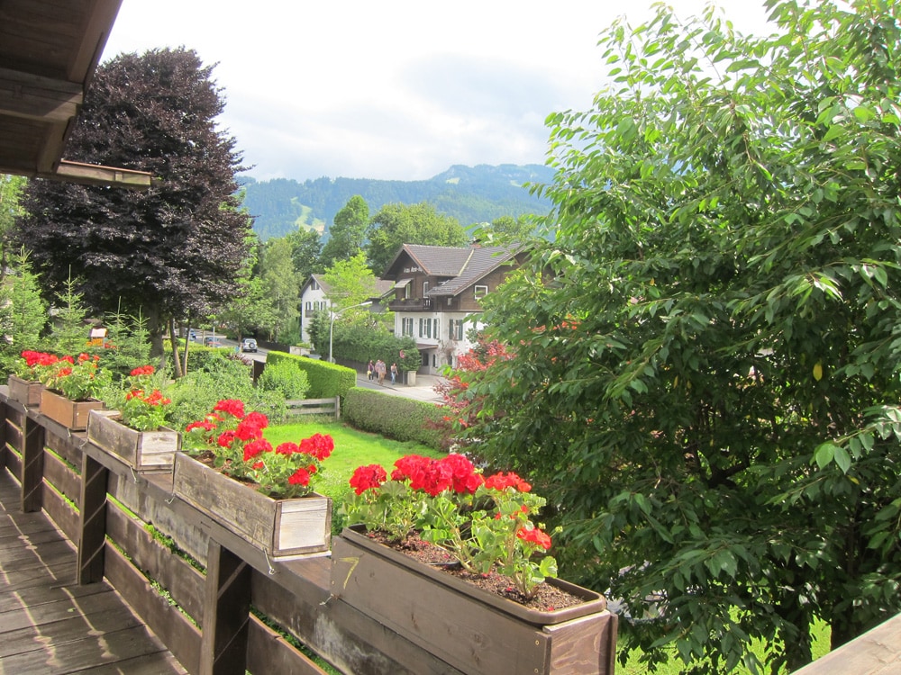 A view from the balcony features vibrant red geraniums in wooden planters, set against a backdrop of lush greenery and distant mountains. The pathway below is surrounded by well-maintained gardens and houses, inviting a peaceful ambiance.