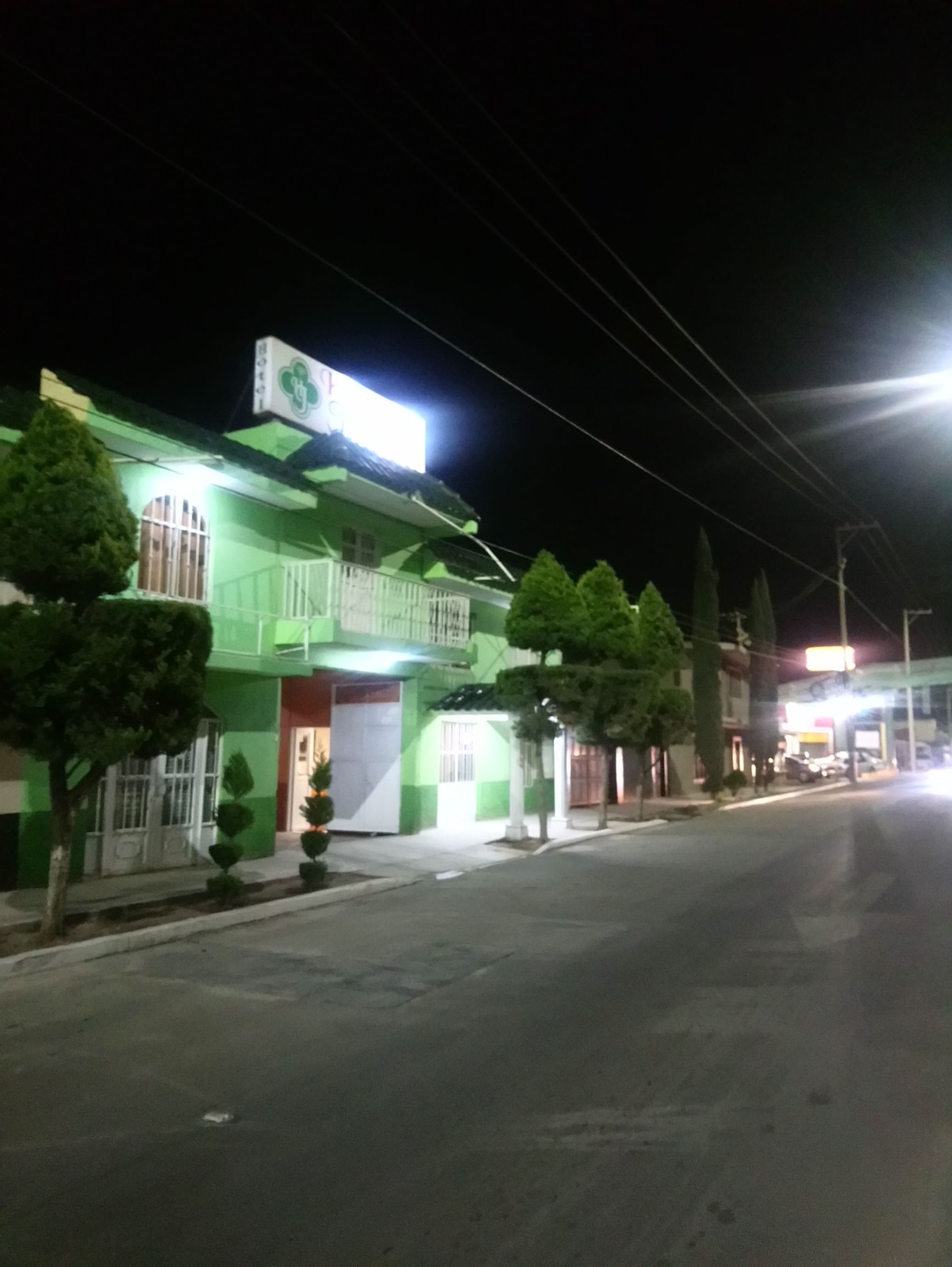 A brightly illuminated building showcases a green facade at night, featuring multiple balconies. Neat, trimmed trees line the street, complementing the structure. Streetlights emit a gentle glow, illuminating the pathway and surrounding area.