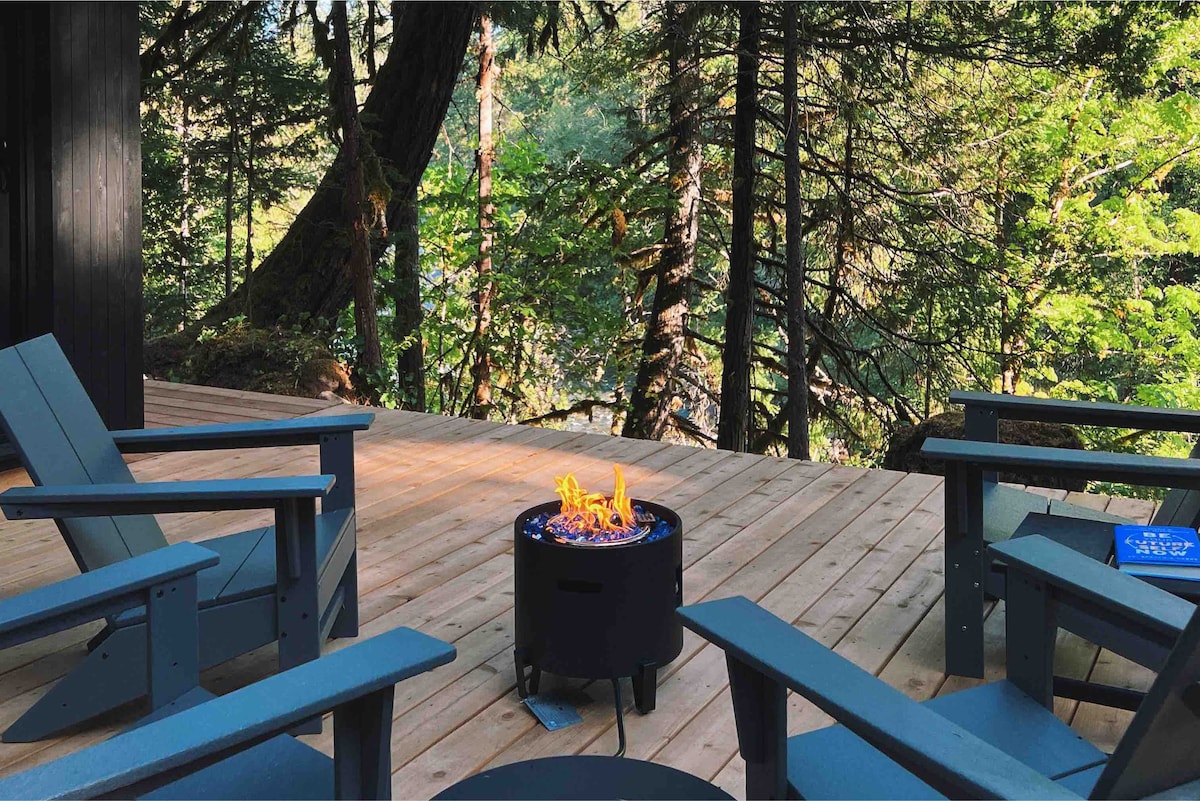 A spacious wooden deck is shown, furnished with several adirondack chairs surrounding a modern propane firepit. Lush green trees and foliage are visible in the background, offering a serene view of nature.
