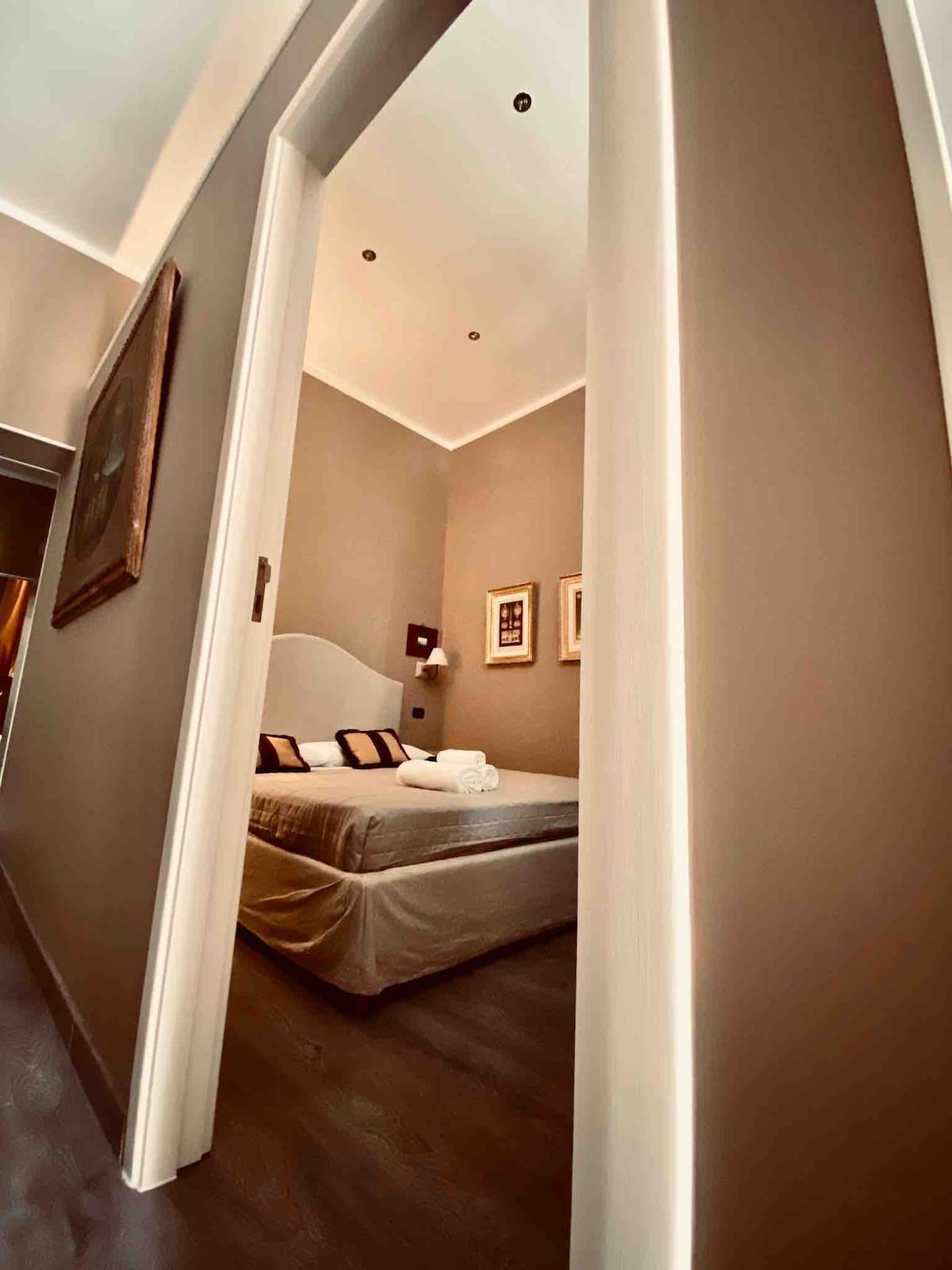 A well-appointed bedroom is glimpsed through an elegantly framed doorway. Soft lighting illuminates the spacious bed, adorned with neutral-toned linens. The walls are painted in a muted shade, complemented by framed artwork, contributing to a serene ambiance.