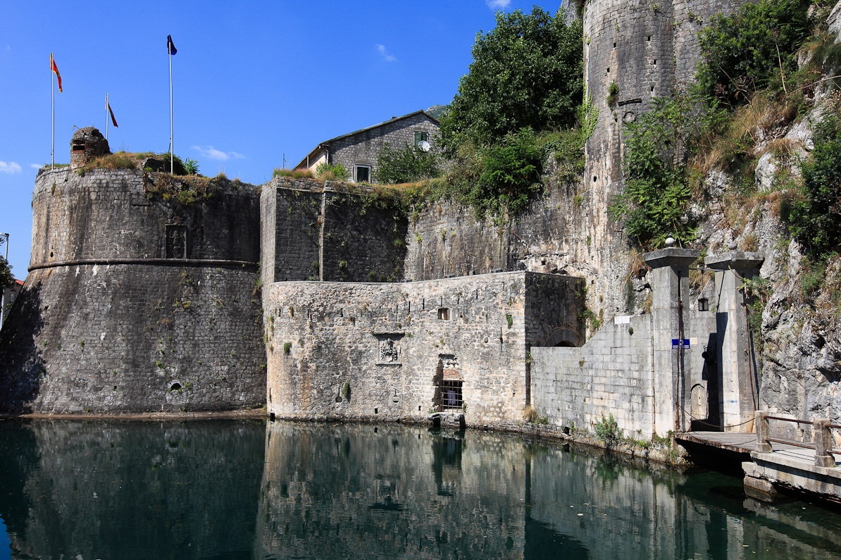 A historic stone fortress is seen rising from the water's edge. It features sturdy walls with scattered greenery and flags gently waving in the breeze. The calm water reflects the structure, enhancing the tranquil atmosphere. The scene is set against a clear blue sky.