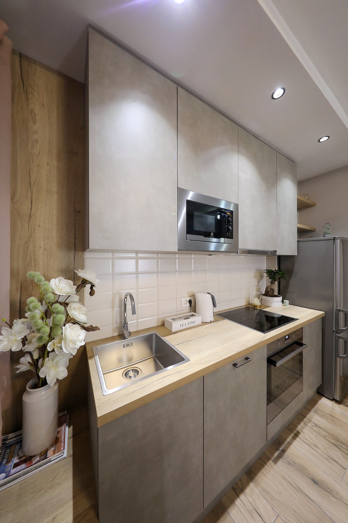 A modern kitchen area features sleek grey cabinetry and a wooden countertop. Stainless steel appliances, including a microwave and refrigerator, are visible. A small sink is located beneath overhead cabinets, with a potted plant and decorative items enhancing the space.