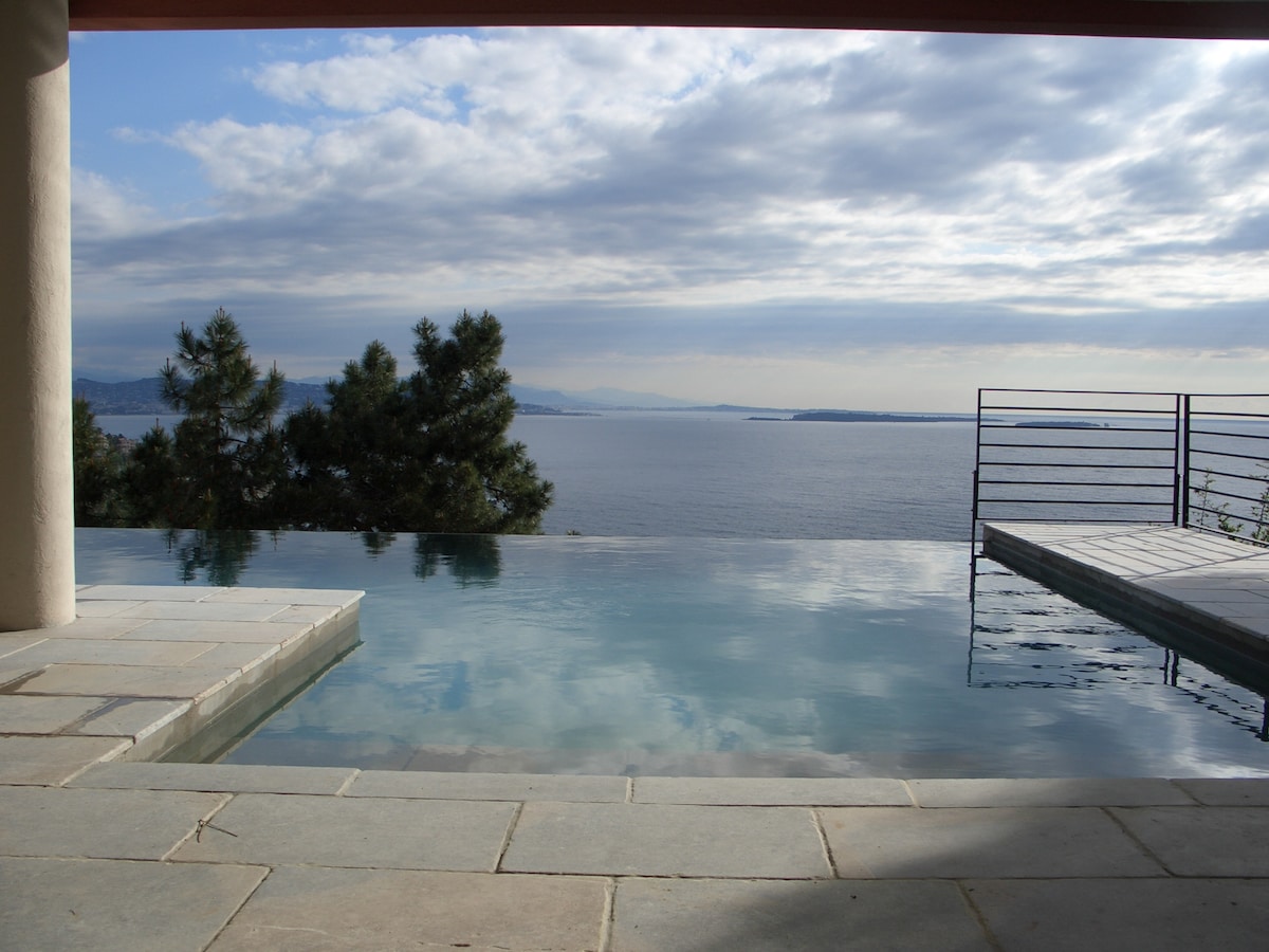 An infinity pool extends towards the sea, reflecting the tranquil waters and sky above. The pool's sleek stone edge blends with the surrounding patio, framed by trees and a stunning view of the coastline and distant islands.