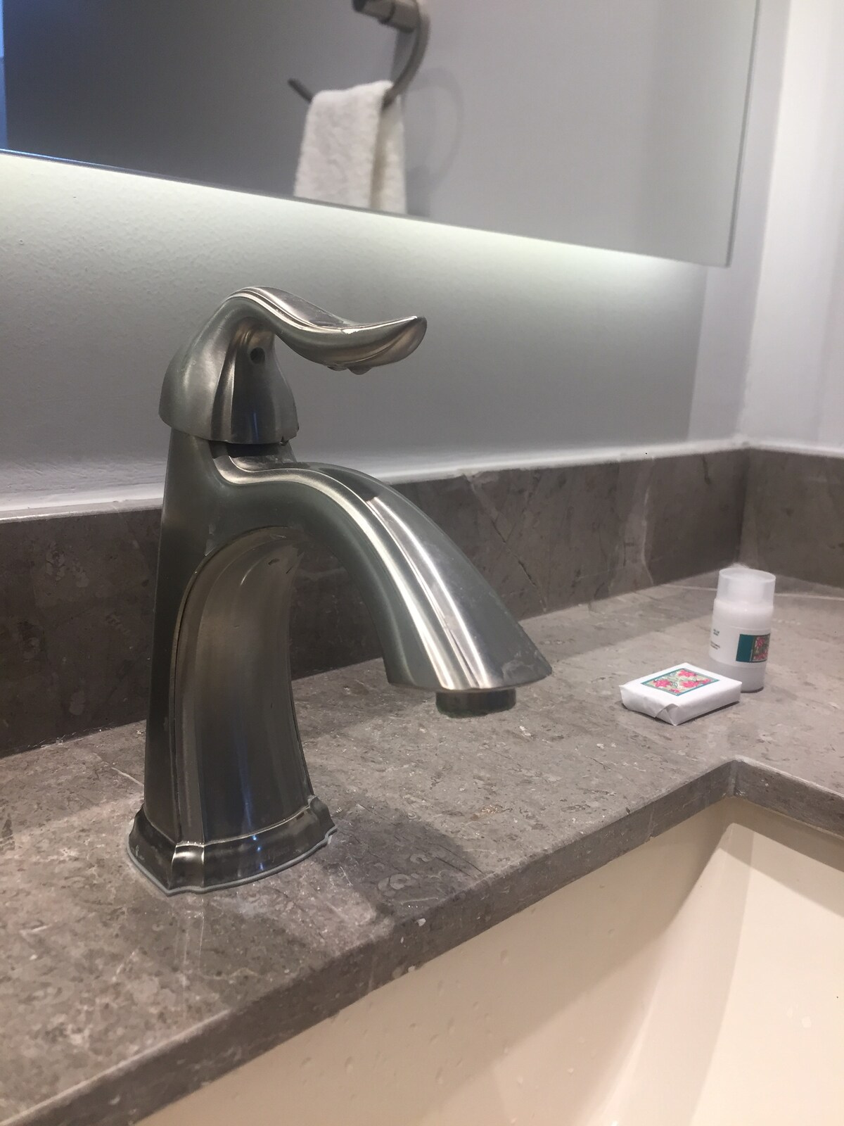 A sleek metal faucet is positioned above a smooth stone countertop, with light reflecting off its polished surface. Nearby, basic toiletries are arranged, including a soap bar and a small bottle, contributing to an uncluttered and practical bathroom setup.