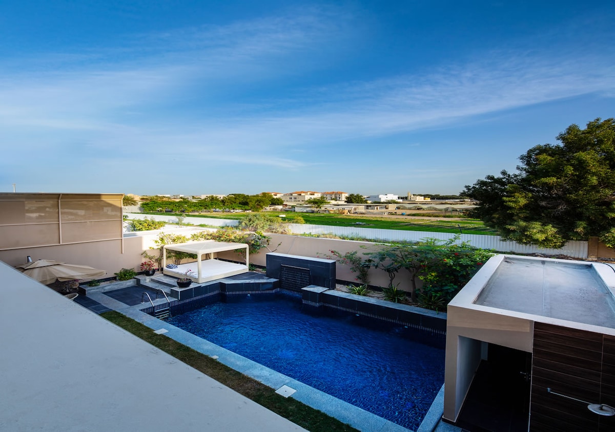 An outdoor space features a private swimming pool surrounded by a stone deck. A covered sitting area offers shade, while lush greenery lines the backdrop. A clear blue sky enhances the relaxing atmosphere of this serene setting.