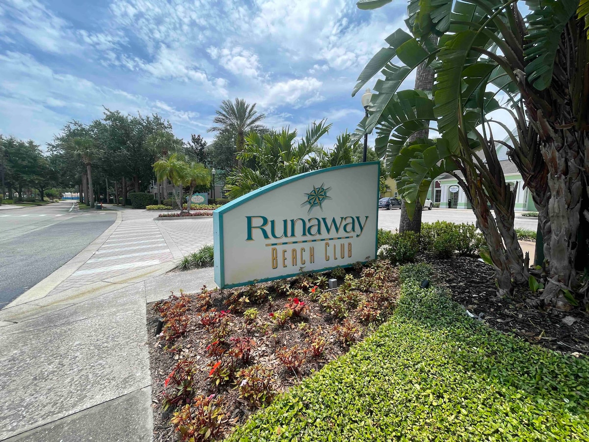 The entrance to Runaway Beach Club is marked by a welcoming sign amidst lush landscaping. Tropical plants and colorful flowers surround the sign, adding a vibrant touch to the well-maintained grounds under a partly cloudy sky.