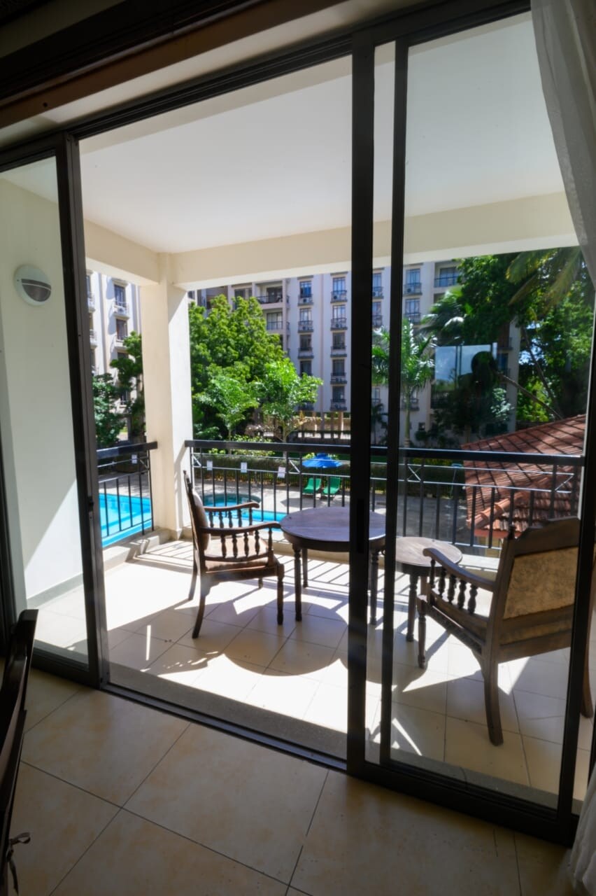 A balcony is visible through sliding glass doors, furnished with two wooden chairs facing a pool area. Green trees and a glimpse of the building's exterior can be seen beyond the balcony, offering a serene outdoor space for relaxation.
