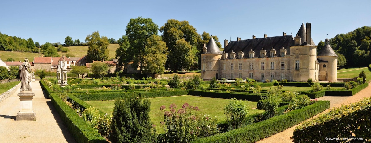 The image features a grand château surrounded by meticulously maintained gardens. Neatly trimmed hedges and vibrant flower beds are visible in the foreground, leading to the elegant stone structure with distinctive turrets. Lush green trees line the distant landscape under a clear blue sky.