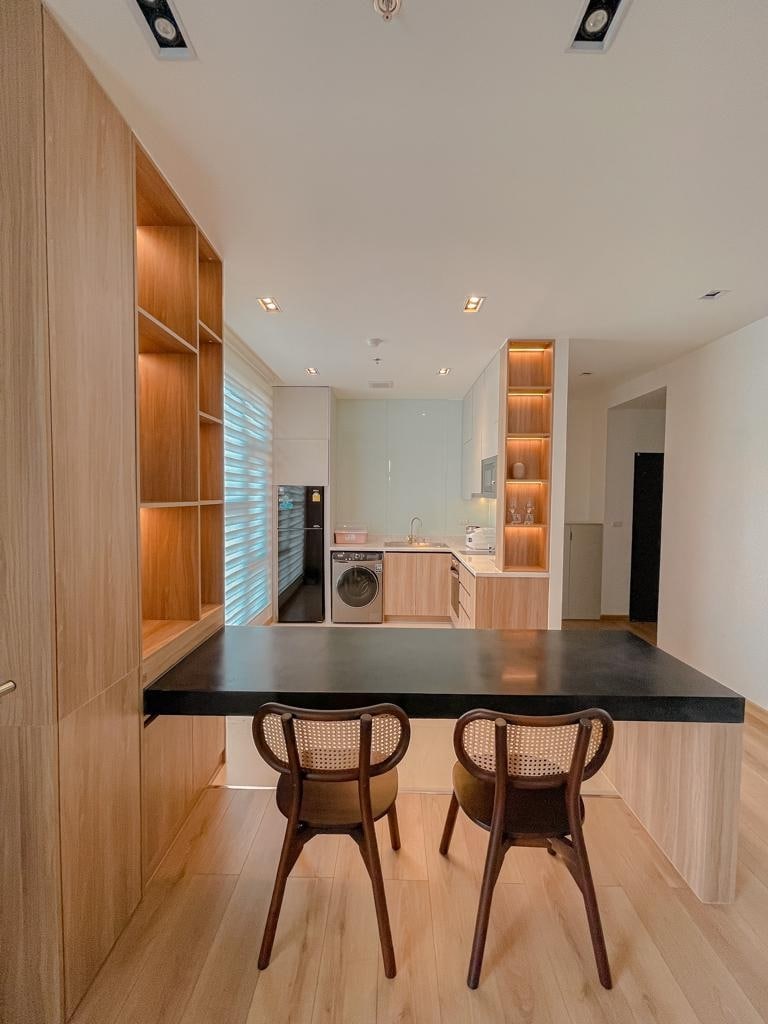 A modern kitchen area is presented, featuring sleek wooden cabinetry and a black countertop. Two stylish woven chairs are positioned at the counter. A washer and dryer are discreetly integrated into the space, with soft lighting enhancing the overall ambiance.