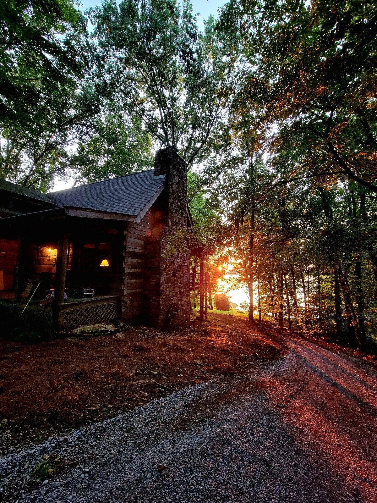 A log cabin exterior is framed by lush trees, with sunlight filtering through the branches. A gravel path leads toward the cabin, while the warm glow of the setting sun is visible in the background, creating a serene ambiance.
