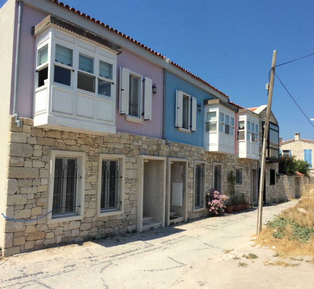 The exterior of a charming building is featured, showcasing a combination of stone walls and pastel-colored facades. Large, light-filled windows are visible, providing ample natural light. The area is bordered by a small pathway and potted plants, enhancing the inviting neighborhood atmosphere.