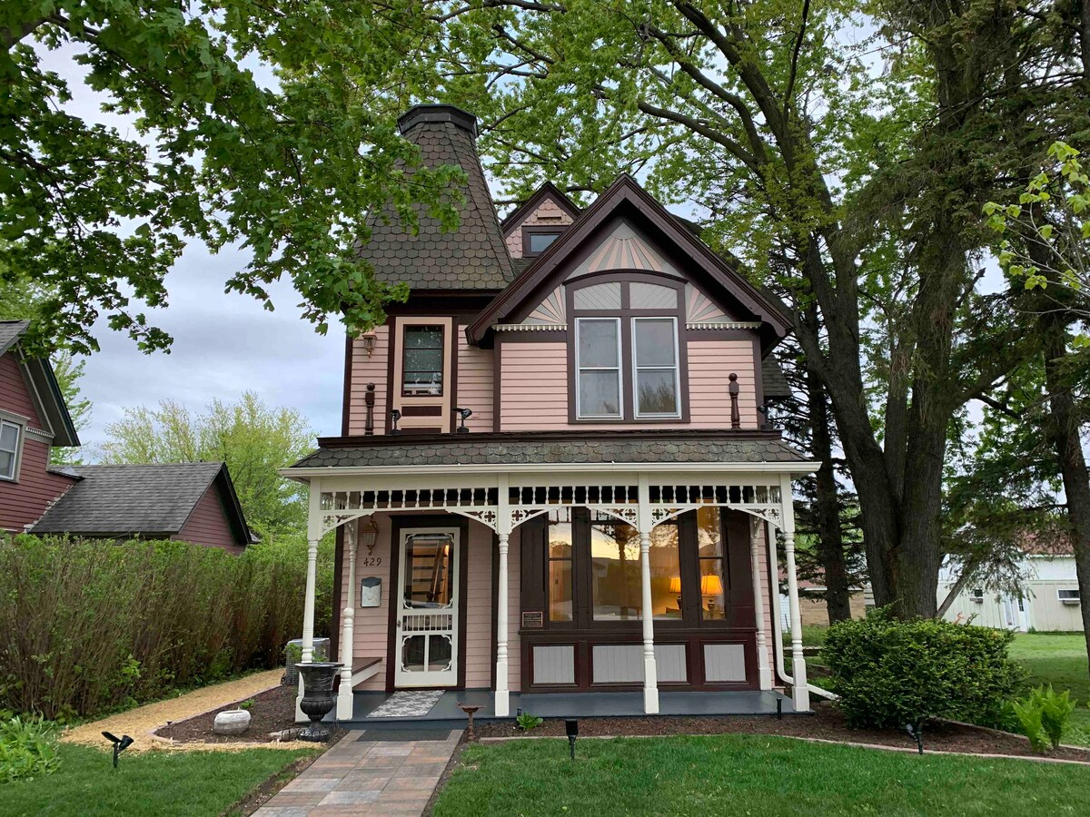 A historic house features a charming exterior with pink siding and intricate architectural details. The front porch is framed by white railings and decorative trim. Lush greenery surrounds the home, and large windows allow natural light into the interior.