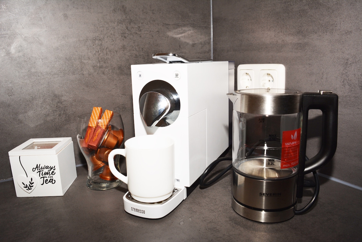 A modern coffee station is displayed, featuring a single-serve coffee maker, a kettle, and a white mug on a saucer. A glass jar filled with chocolate treats sits nearby, alongside various coffee capsules arranged for easy access.