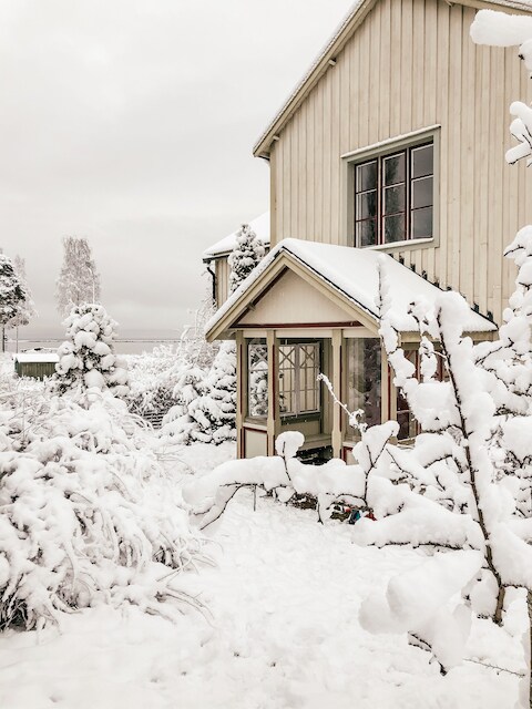 A charming house is partially visible, framed by snow-covered trees and bushes. The entrance features a glass-paned door and a covered porch, with intricate wooden details complementing the tranquil winter scene.