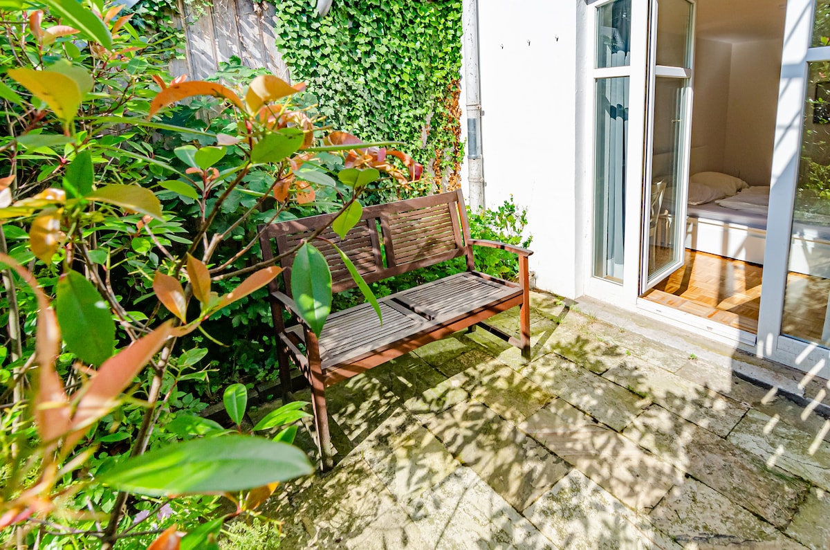 A wooden bench is positioned on a stone patio, surrounded by lush greenery. Sunlight casts soft shadows on the ground, highlighting the serene outdoor space. The entrance to the apartment is visible in the background, offering access to a peaceful retreat.