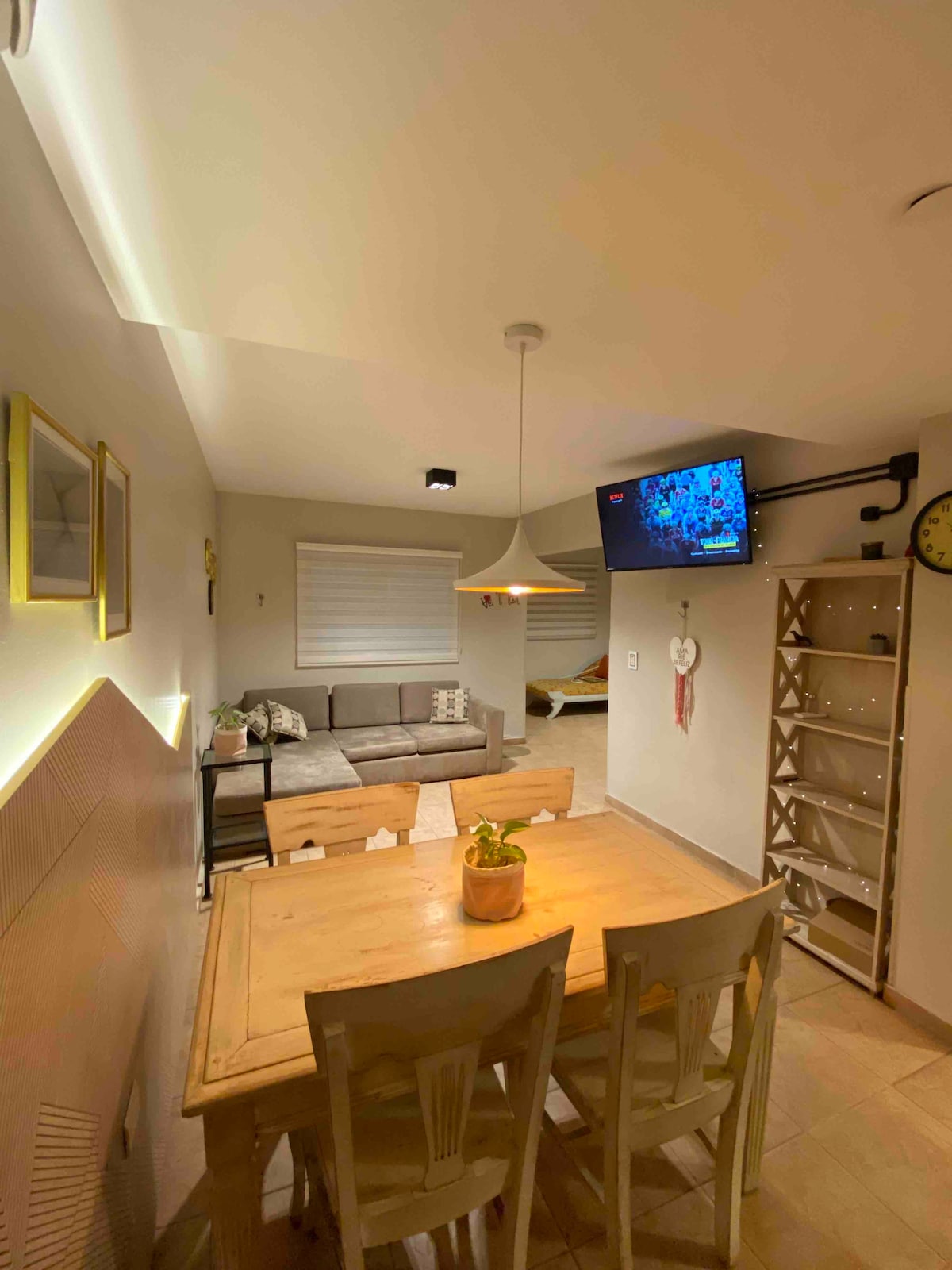 A dining area is visible, featuring a wooden table surrounded by four chairs. A light fixture hangs above the table, and a flat-screen television is mounted on the wall. A comfortable sofa is positioned in the corner, with natural light filtering through a window covered by shades.