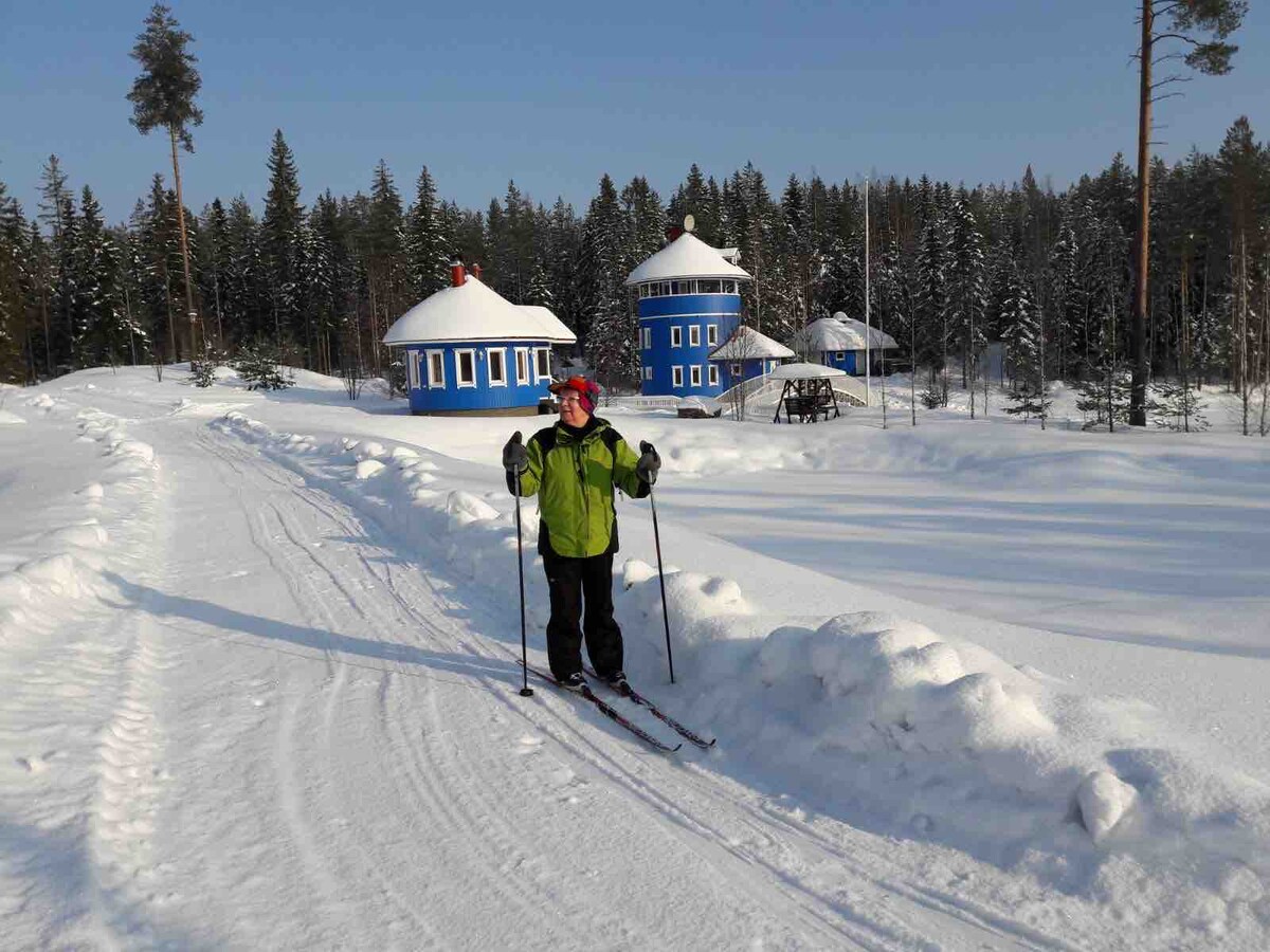 A skier is positioned along a snow-covered path, surrounded by a serene winter landscape. Two blue structures can be seen in the background, framed by evergreen trees dusted with snow. The scene conveys a peaceful, wintry atmosphere.